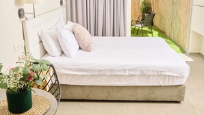 A bed with white linens, a green potted plant, and a chair in a room with a view of a grassy area through a bamboo fence. - Hotel H7 tlv (Tel Aviv)