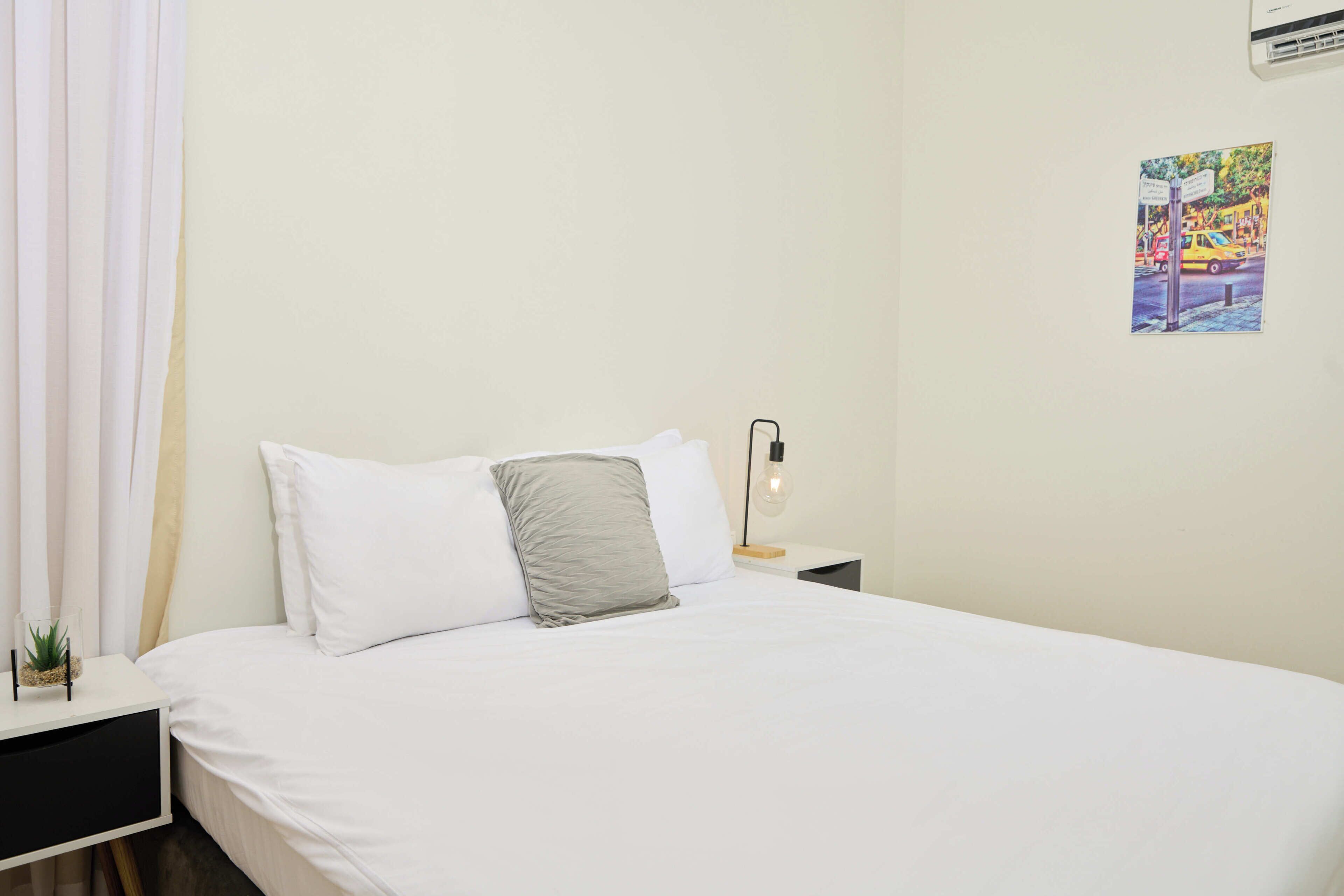 A neatly made bed with white and gray pillows, a bedside table with a lamp, and a wall-mounted air conditioner.