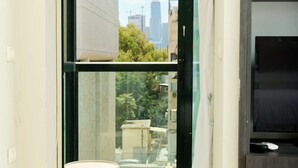 A room with a sliding glass door leading to a balcony, a dark wooden cabinet, and two round ottomans. - Hotel H7 tlv (Tel Aviv)