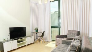 A modern living room with a flat-screen TV, a sofa, a small table with a chair, and a large window with curtains. - Hotel H7 tlv (Tel Aviv)