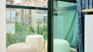 A balcony with two round ottomans, a glass door, and a view of buildings and greenery. - Hotel H7 tlv (Tel Aviv)
