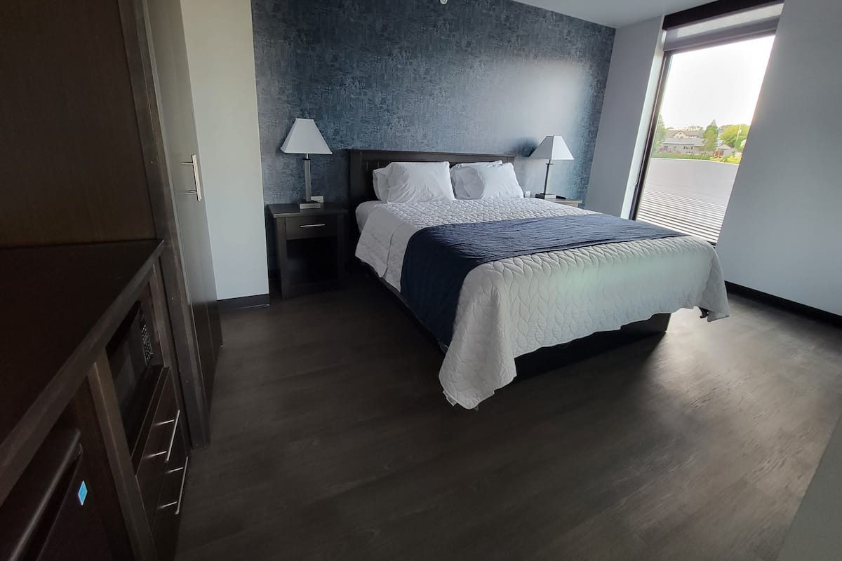 A modern bedroom with a large bed, two bedside tables with lamps, a dark wooden wardrobe, and a view of the outside through a large window.