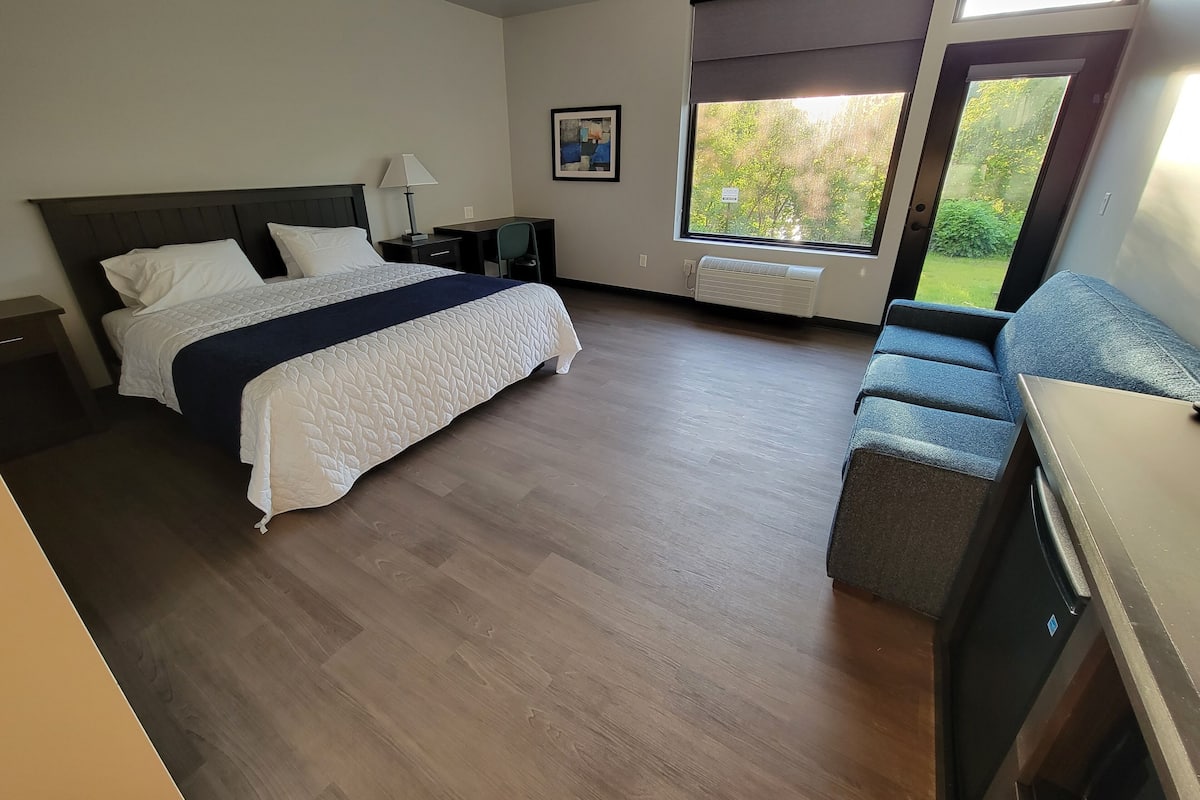 A modern hotel room with a large bed, a desk, a chair, and a window with a view of greenery.