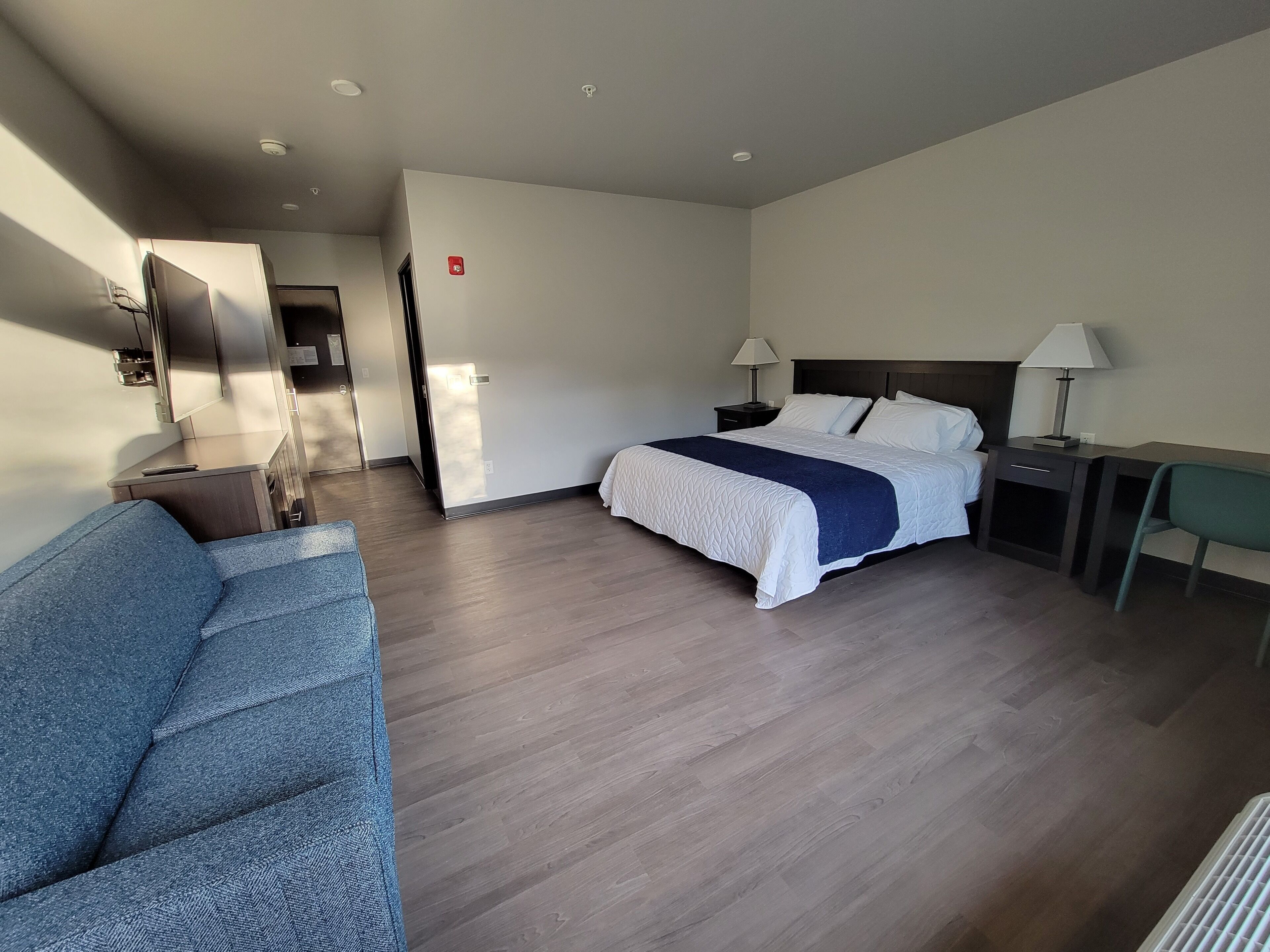 Habitación de hotel con cama, escritorio, silla, sofá y baño visible a través de una puerta abierta.