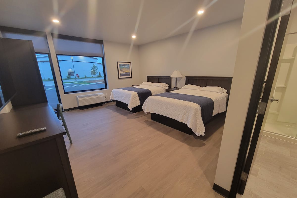 A hotel room with two beds, a desk with a remote control, a window with a view of the street, and a door leading to a shower area.