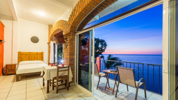 A balcony with a view of the sea, a bed, a table with chairs, and a clock on the wall. - Traditional Sierra Leon Oceanfront Rooms (Puerto Vallarta)