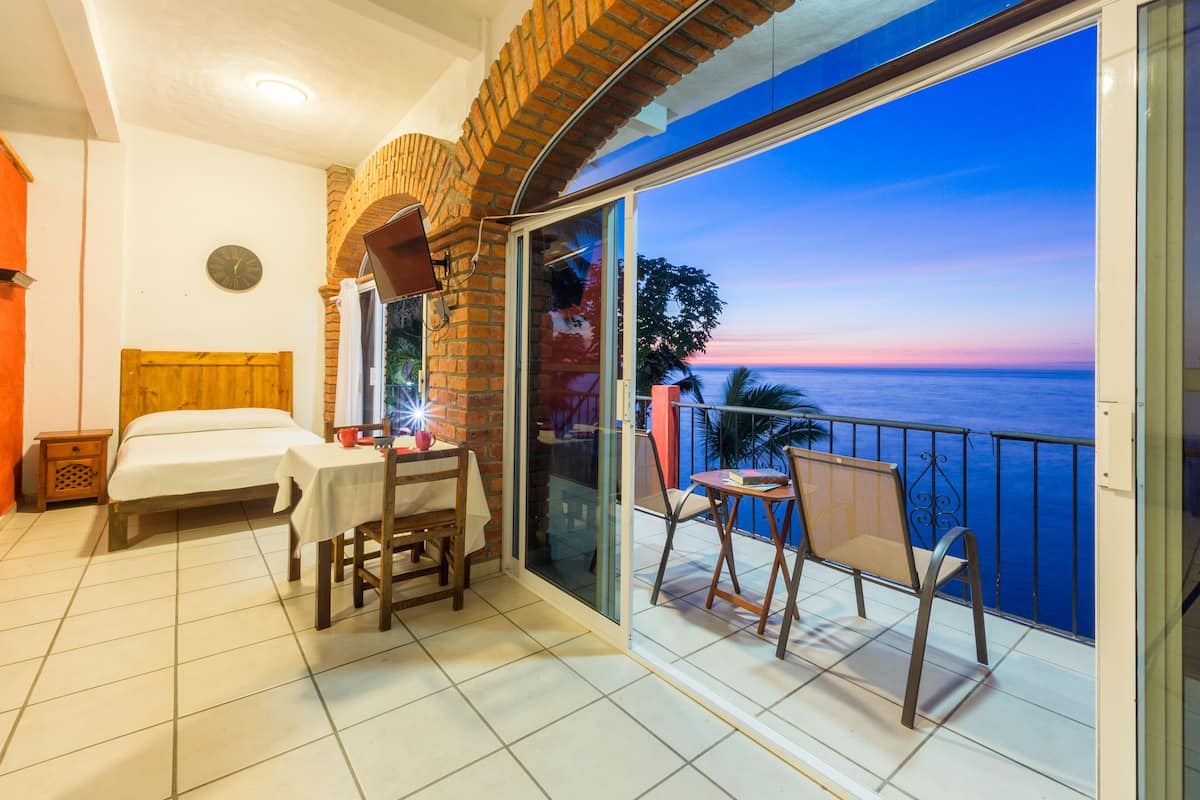Un balcón con vista al mar, una cama, una mesa con sillas y un reloj en la pared.
