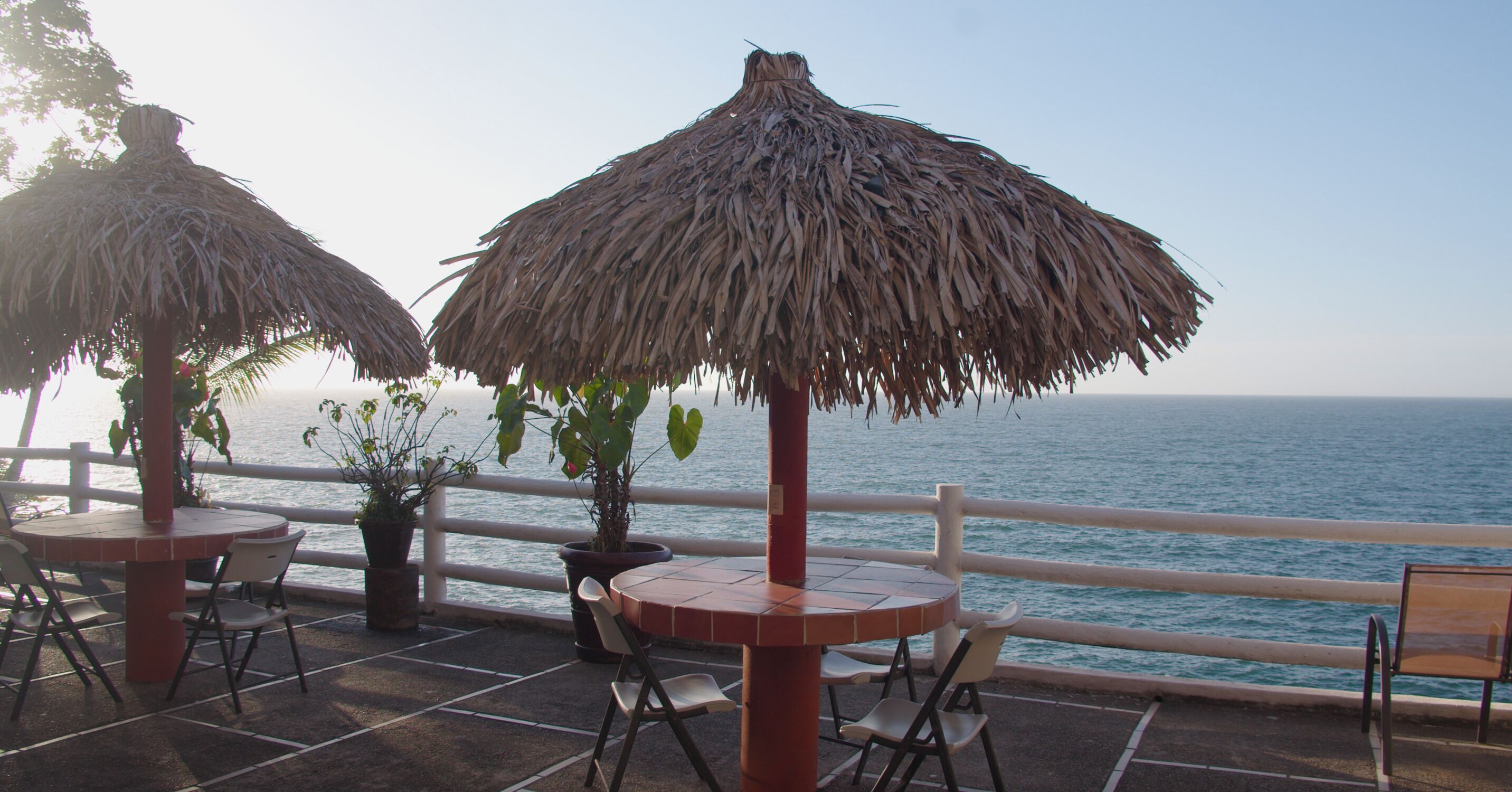 Un patio frente al mar con mesas y sillas bajo sombrillas de paja, con vista al océano.