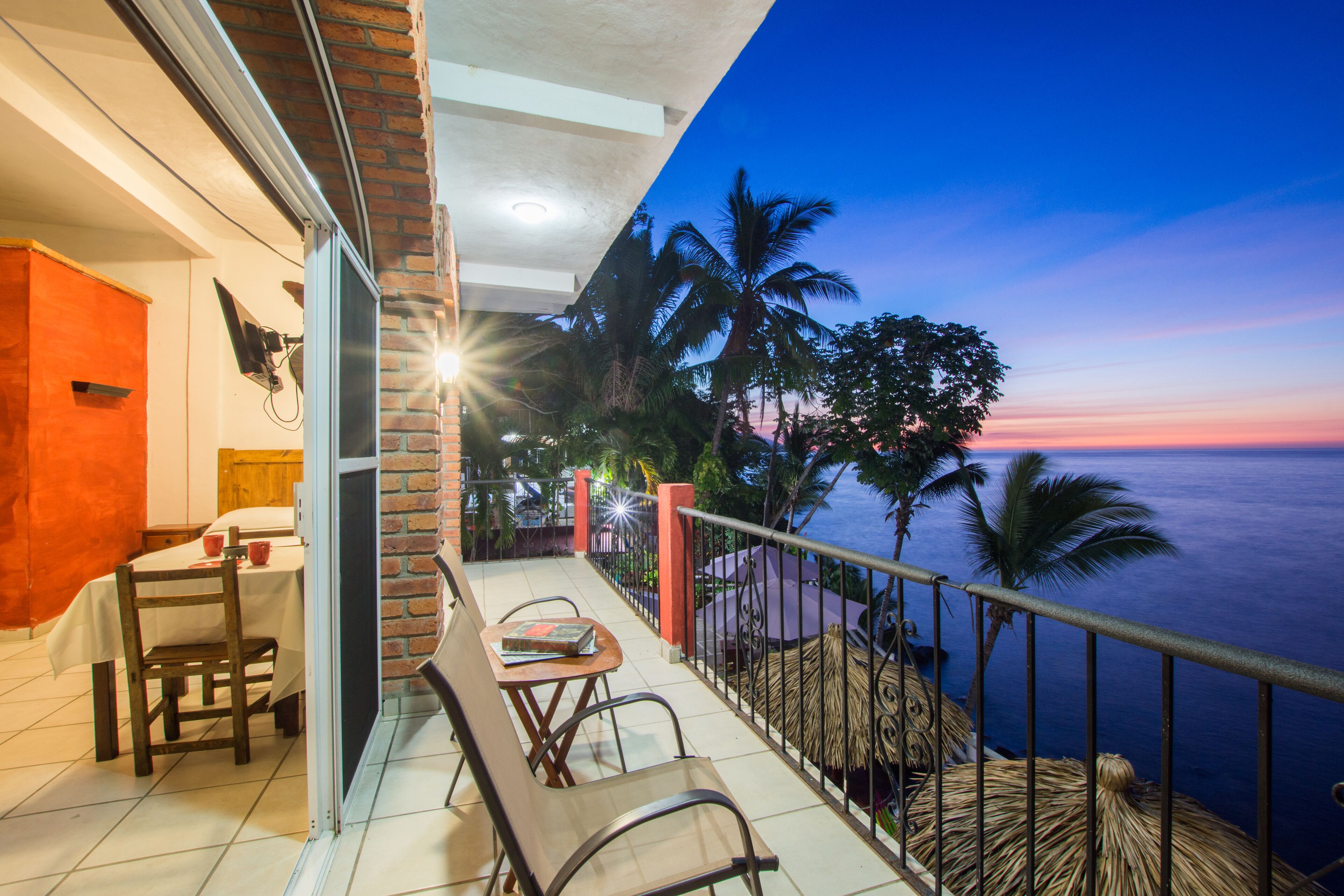 A balcony with a table and chairs, overlooking a body of water and palm trees at dusk.