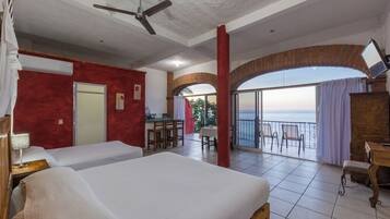 A hotel room with two beds, a balcony with a view of the sea, a dining area, and a TV.