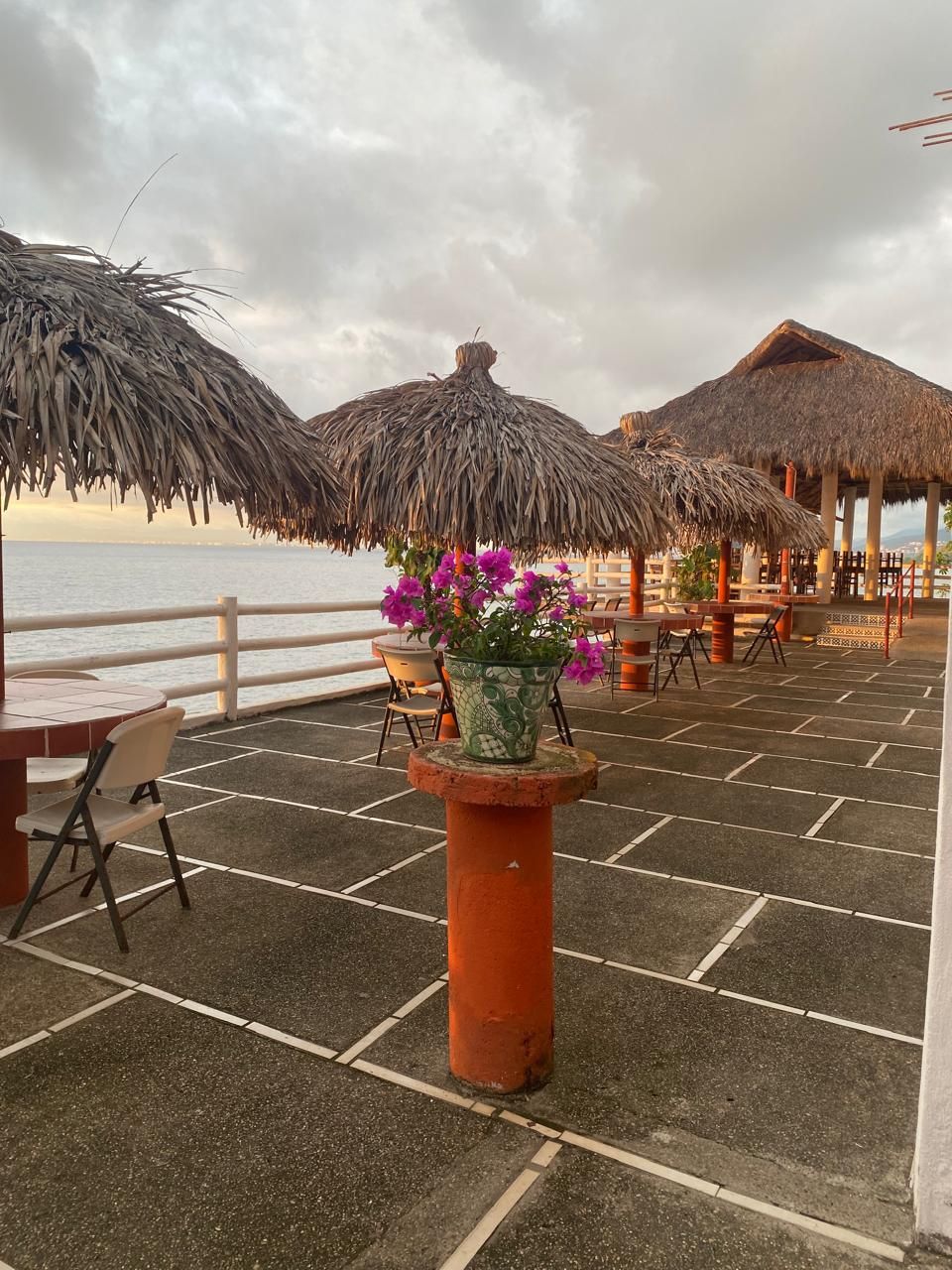 Un área de estar al aire libre con sombrillas de paja, una mesa con sillas y una planta en maceta con flores moradas.