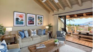 Una sala de estar con sofá, mesa de centro y cuadros en la pared. Hay un balcón con mobiliario exterior y vistas al océano.