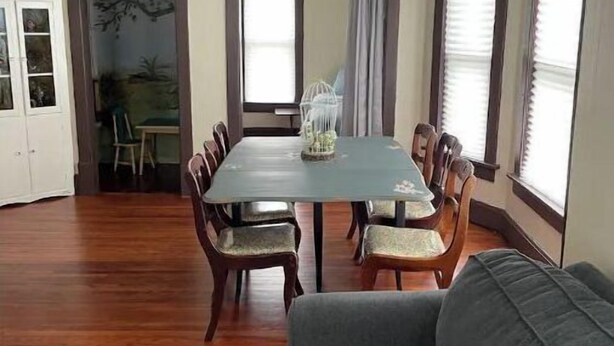 A dining area with a rectangular table surrounded by wooden chairs, a gray sofa, and a patterned rug. - Fairy Bowery (Nauvoo)