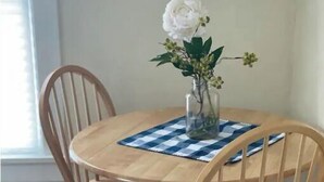 A round wooden dining table with a checkered tablecloth, two wooden chairs, and a vase with a white flower. - Fairy Bowery (Nauvoo)