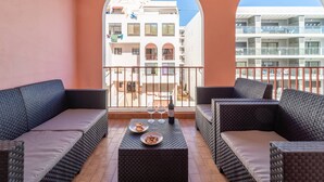 A balcony with wicker furniture, a table with a bottle and glasses, and a view of buildings. - Quarteira Beach - 2.º D by HelloVacations (Loulé)