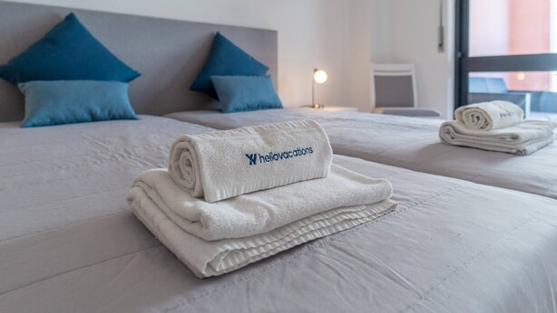 A hotel room with two beds, each with a neatly folded white towel and a blue pillow. - Quarteira Beach - 2.º D by HelloVacations (Loulé)