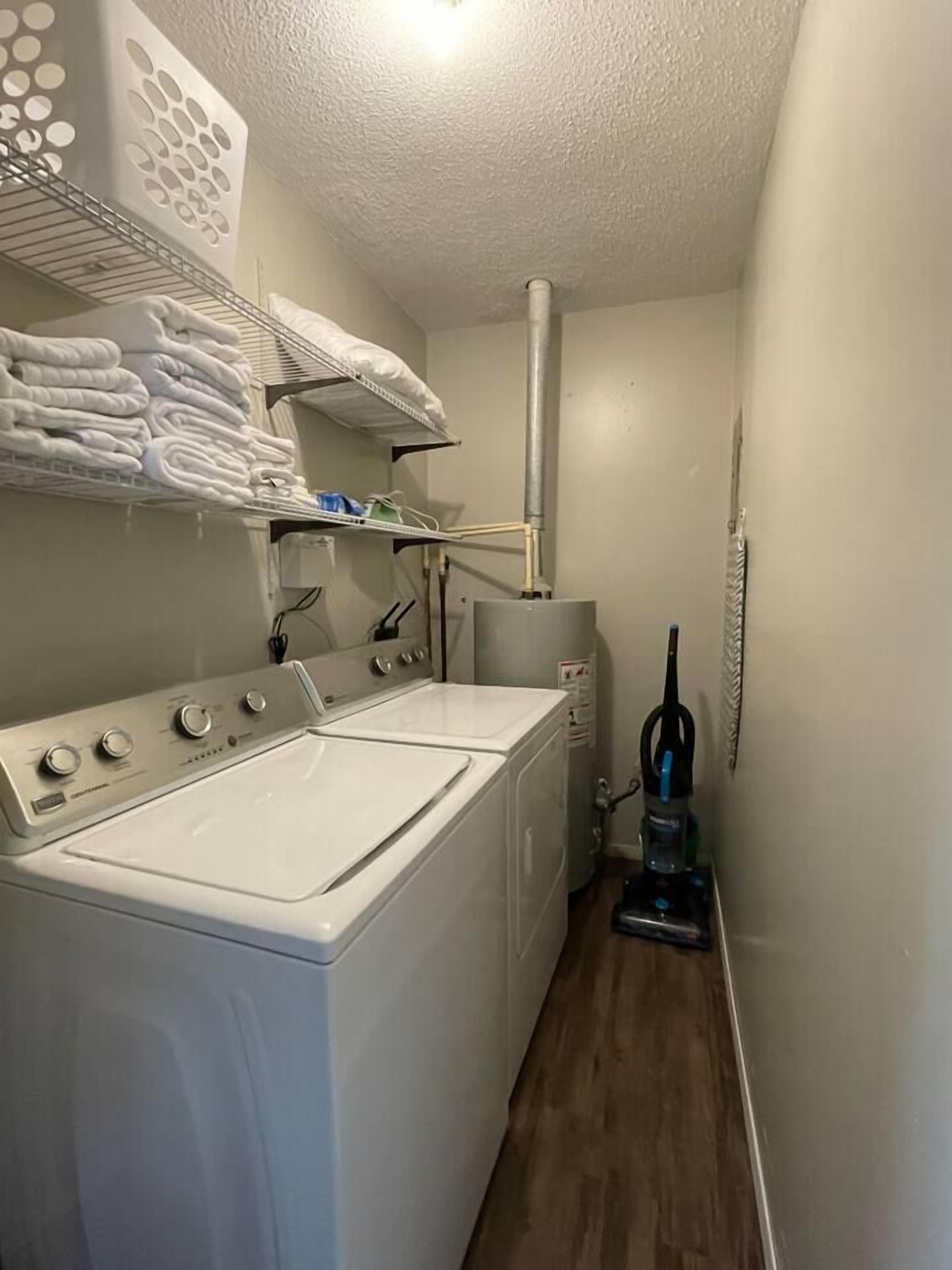 Un cuarto de lavandería con lavadora y secadora de carga frontal, un estante con toallas dobladas y una aspiradora.