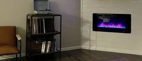 A living room with a flat-screen TV mounted above a fireplace, a bookshelf, and a coffee table with a mug.