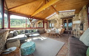 A spacious room with a stone wall, wooden ceiling, and large windows offering a view of the countryside.