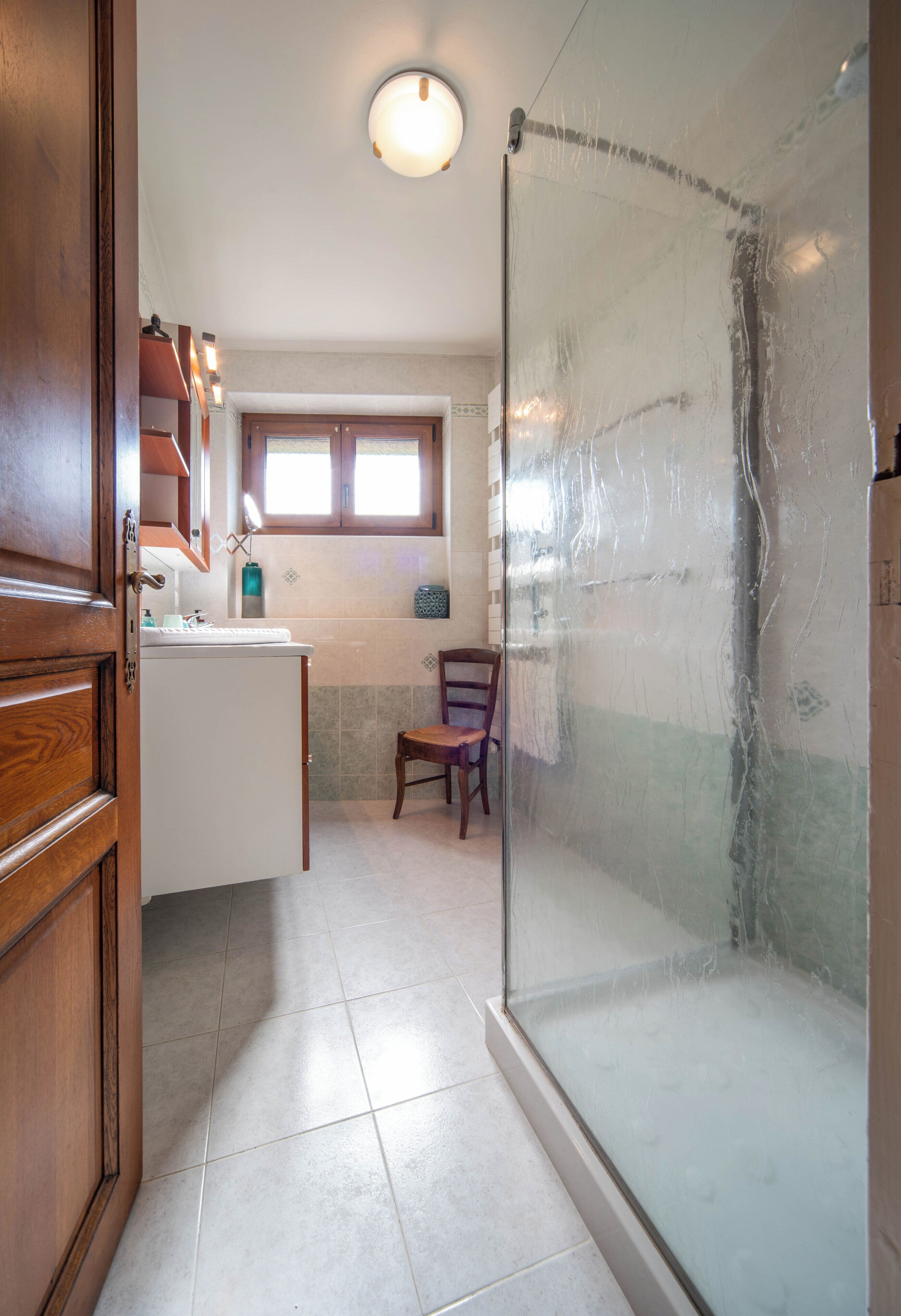 A bathroom with a glass shower enclosure, a wooden door, a sink with a mirror, and a window.