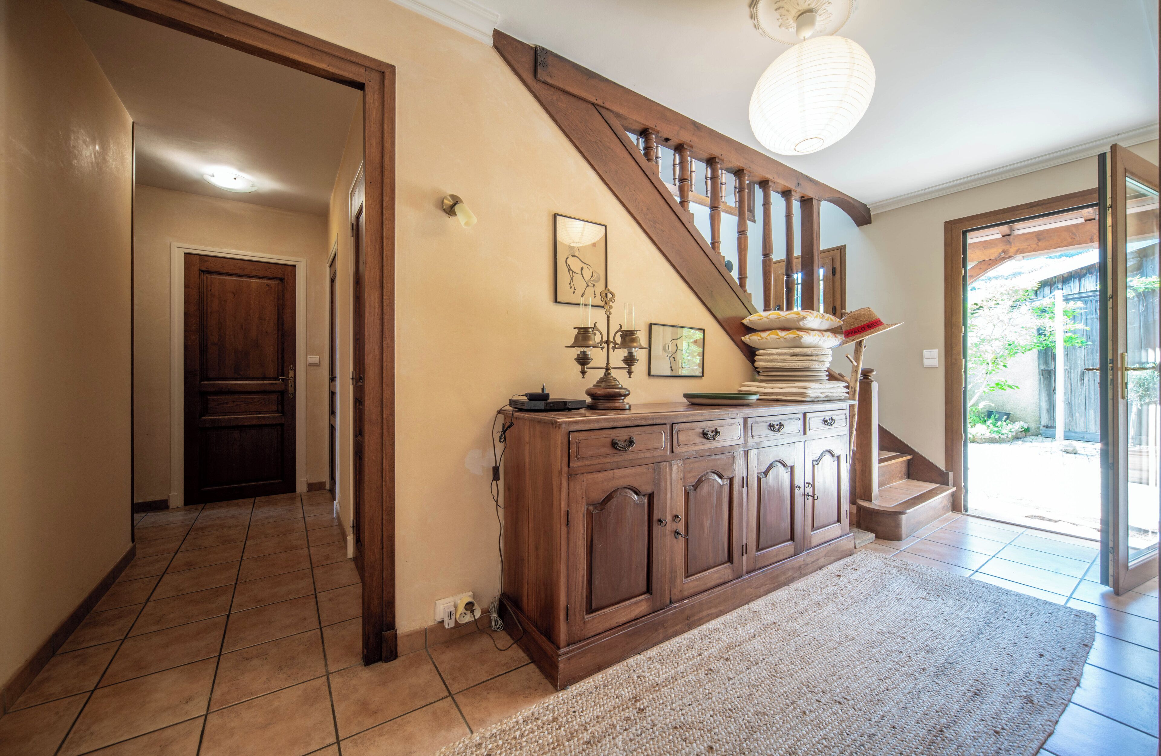 Un couloir avec un escalier en bois, une armoire contenant des objets décoratifs et une porte donnant sur une autre pièce.