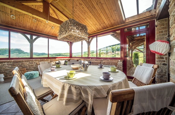 A wooden dining area with a table set for four, large windows, and a view of the countryside.