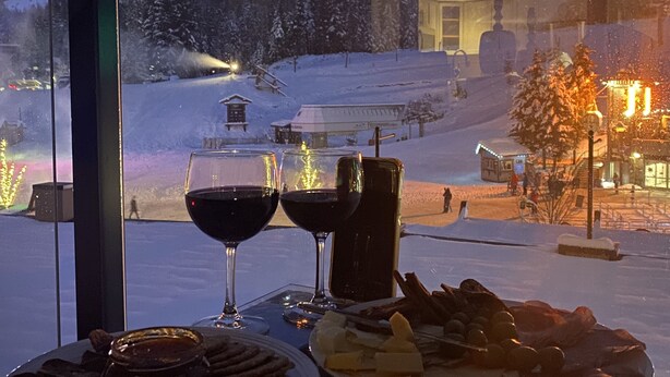 A table with a plate of food and two glasses of red wine, a bottle, and a glass of water, with a snowy mountain view through a window. - BEST Location! Stunning Mountain Views, Ski-in/Ski-out, Base of both lifts!⛷🚠 (Whistler)