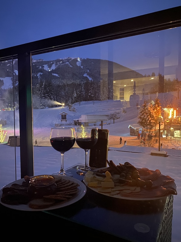 Mesa com prato de comida e dois copos de vinho tinto, uma garrafa e um copo de água, com vista para uma montanha nevada através de uma janela.
