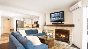A modern living room with a fireplace, a television mounted above it, and a kitchen in the background. - BEST Location! Stunning Mountain Views, Ski-in/Ski-out, Base of both lifts!⛷🚠 (Whistler)
