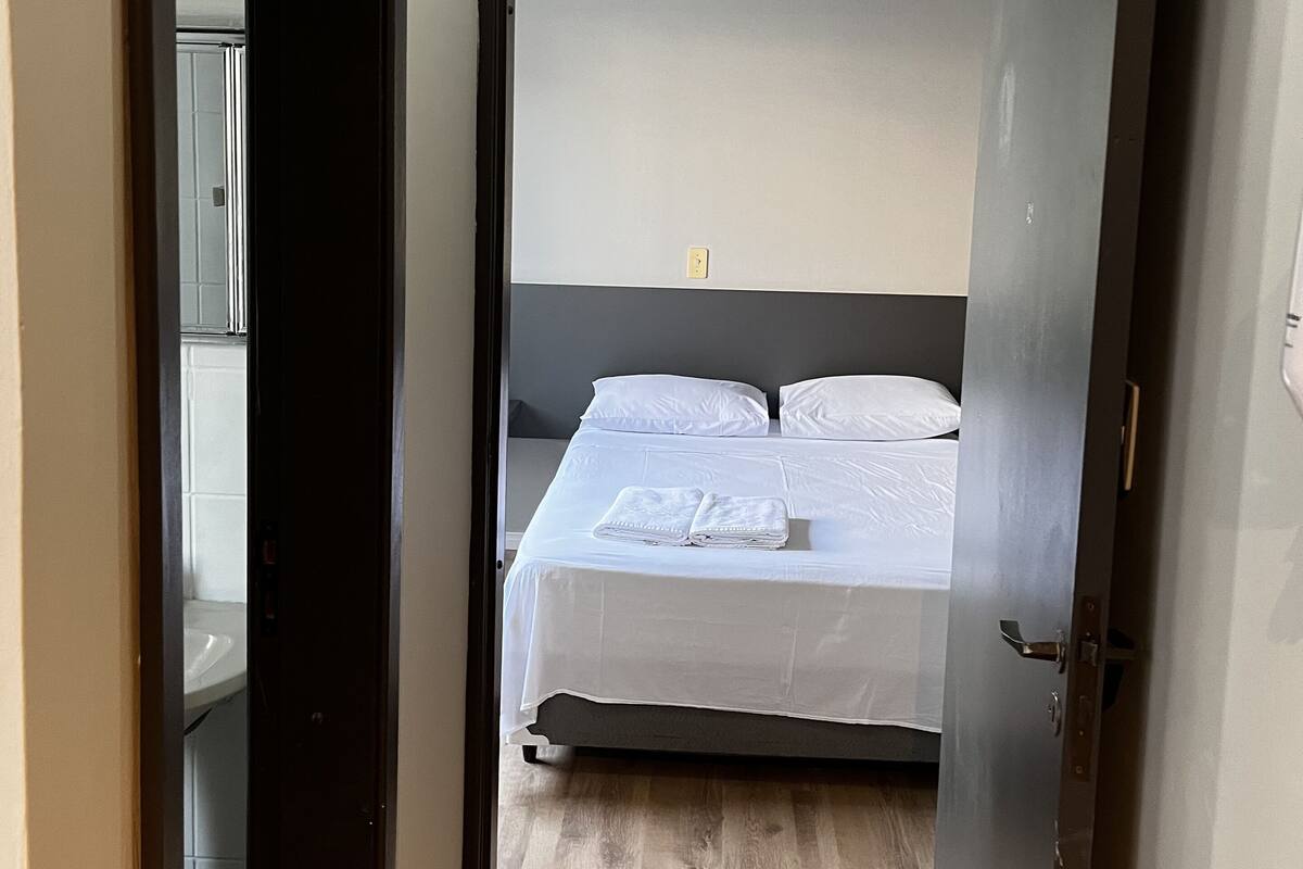 A narrow hallway leading to a bedroom with a bed, a wooden floor, and a white wall.