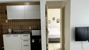 Una cocina compacta con armarios blancos, un lavamanos y un refrigerador pequeño. Un televisor está montado en la pared en la habitación contigua.