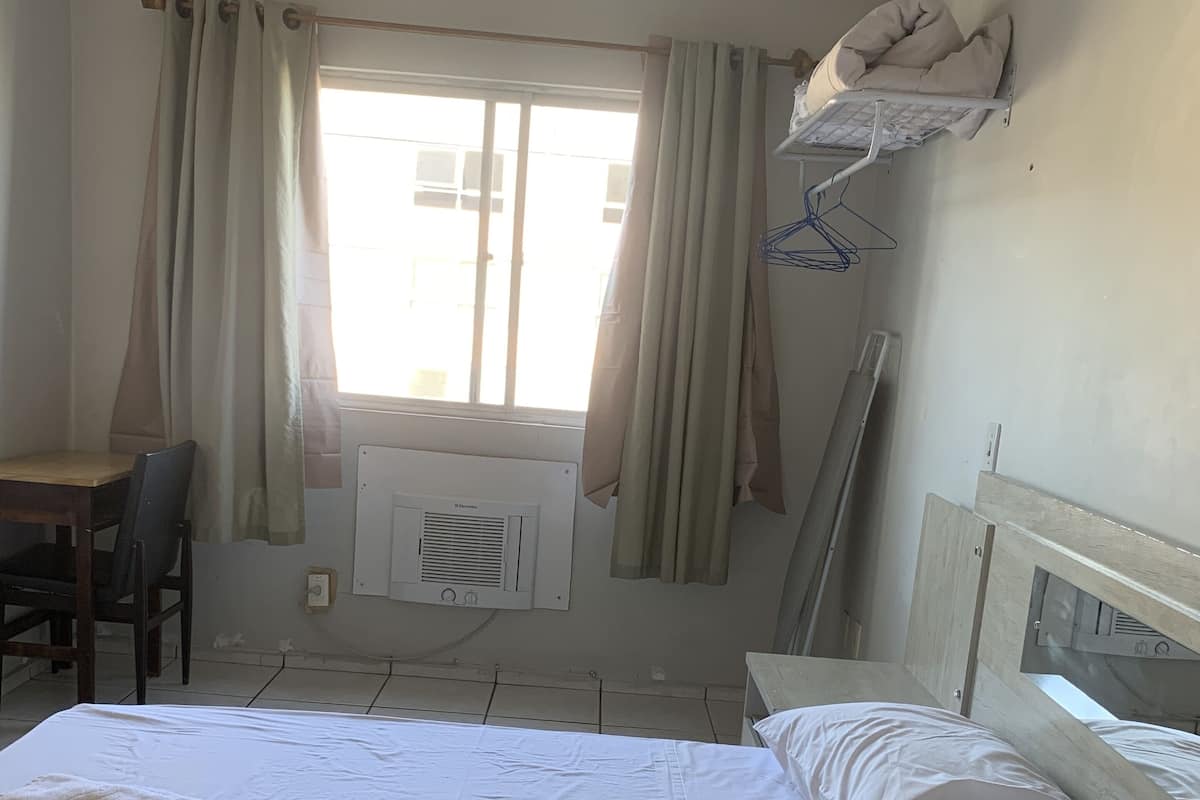 A hotel room with a bed, a desk, a chair, a radiator, and a window with curtains.