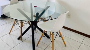 Un comedor moderno con una mesa de vidrio y sillas blancas, un sofá blanco con un cojín gris, y tres obras de arte con hojas enmarcadas en la pared.