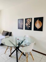 Uma sala de jantar moderna com uma mesa de vidro e cadeiras brancas, um sofá branco com almofada cinza, e três obras de arte com folhas emolduradas na parede.