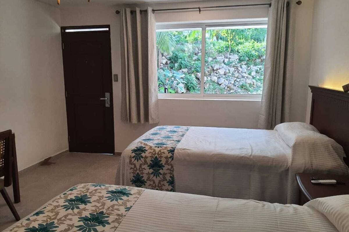 Ein Hotelzimmer mit zwei Betten, einem Fenster mit Vorhängen, einer Tür und einem kleinen Tisch mit Fernbedienung.