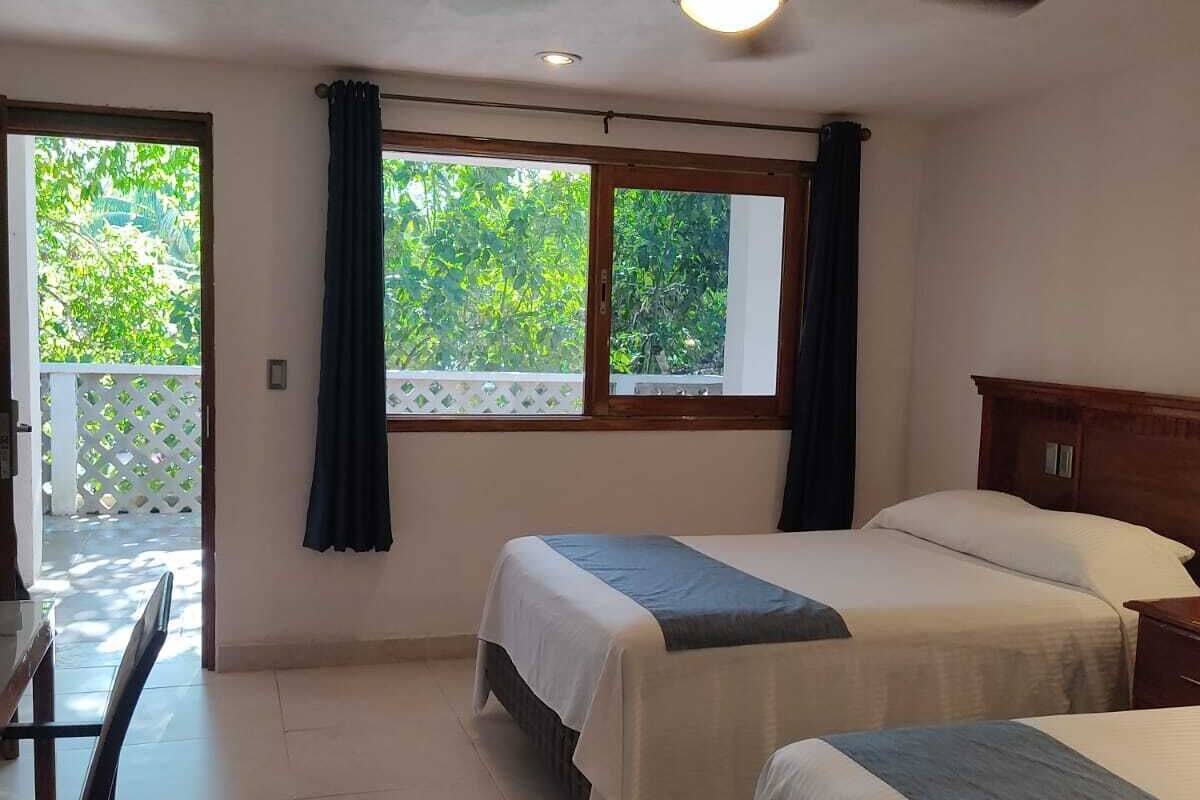 Ein Hotelzimmer mit zwei Betten, einem Deckenventilator, einem Schreibtisch und Blick auf Grün hinter den Fenstern.