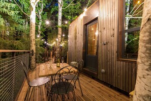 An outdoor wooden deck with a table and chairs, surrounded by trees and string lights.