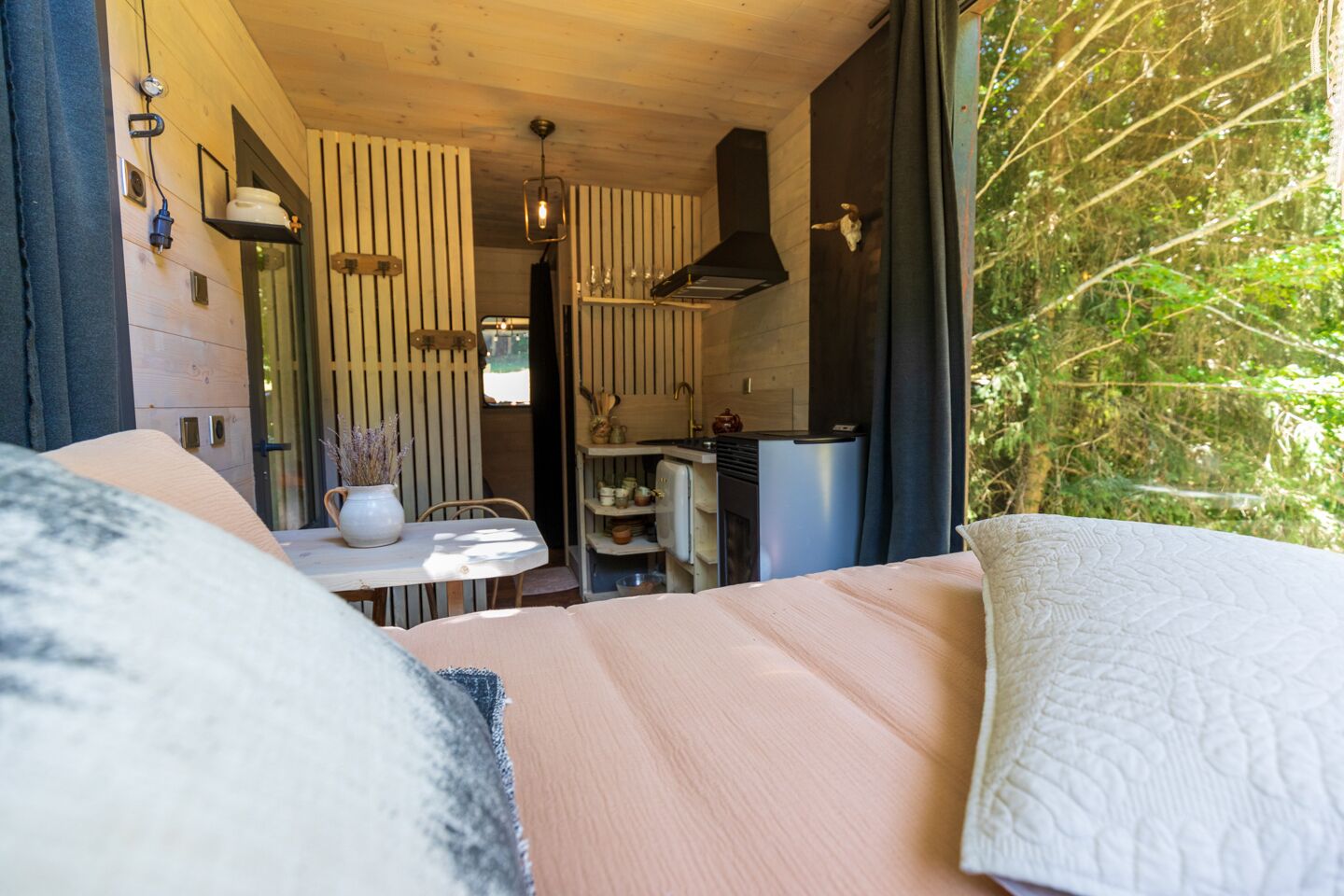 Une chambre à coucher avec un lit, une petite table sur laquelle est posé un vase, et une vue sur une cuisine et un espace extérieur.