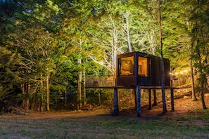 A treehouse with a glass window and a wooden deck, surrounded by dense forest.