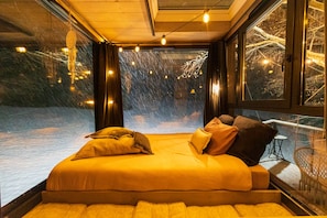 A bedroom with a large bed, a view of the outdoors through a window, and a balcony with a chair.