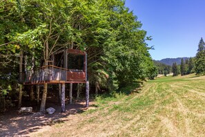 Una estructura rústica de casa en los árboles construida sobre pilotes, rodeada de un bosque frondoso y un cielo despejado.