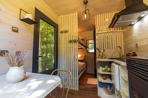 A compact cabin interior with a dining area, a kitchenette, and a sleeping area with a window overlooking greenery.