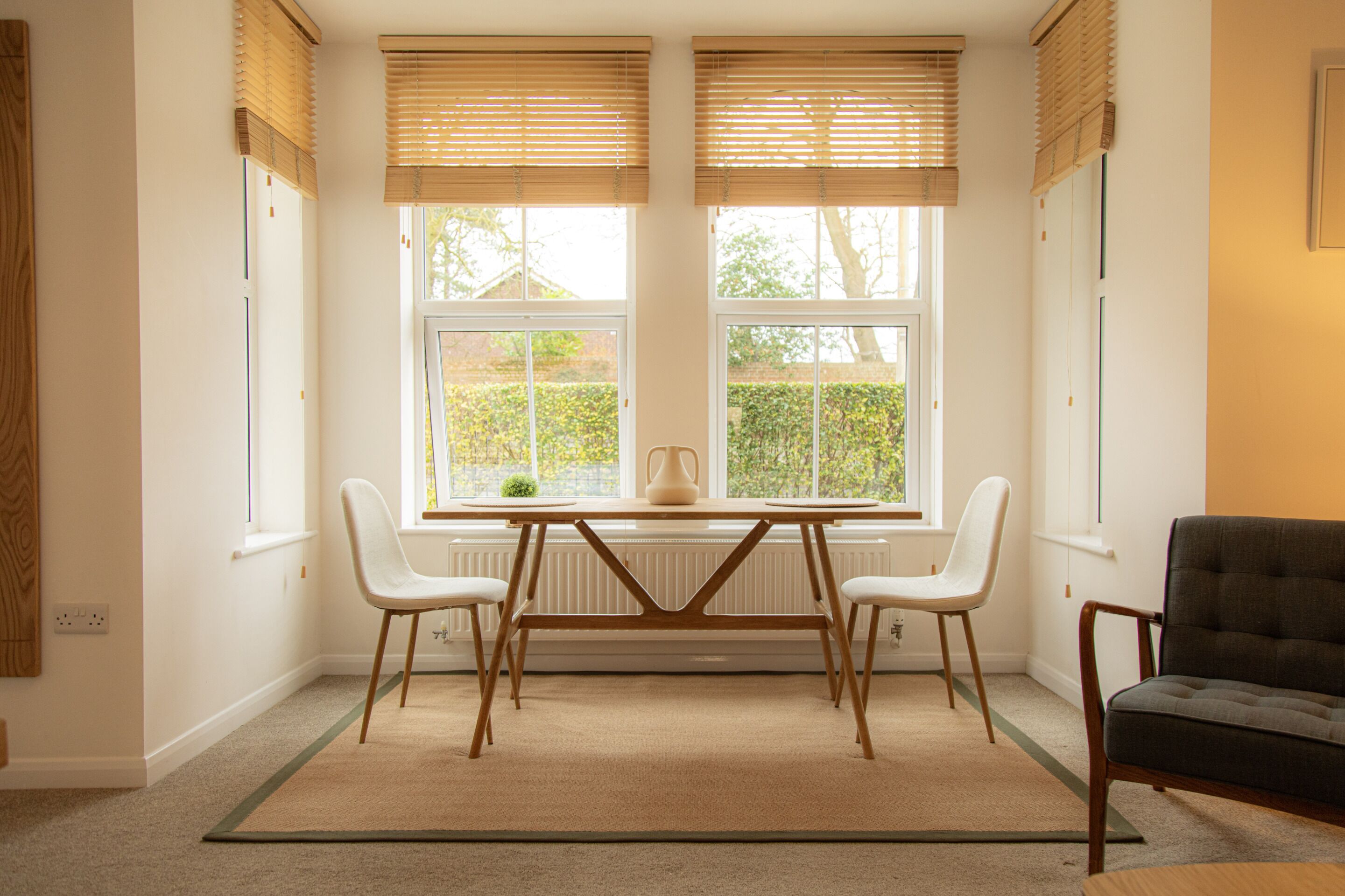 A room with a table and chairs, large windows with blinds, a radiator, and a dark armchair.