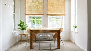 Een eethoek met een houten tafel, vier stoelen en twee ramen met jaloezieën.