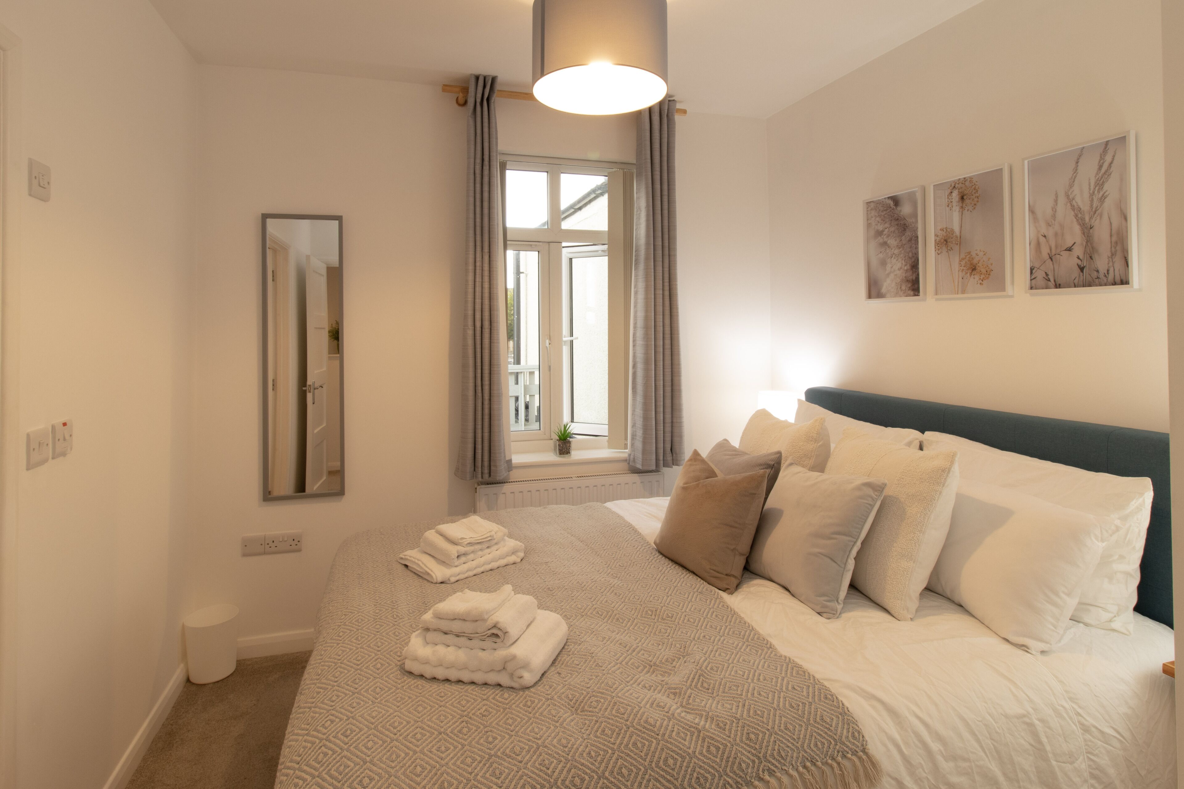 A neatly arranged bedroom with a large bed, a bedside table, a mirror, and a window with curtains.