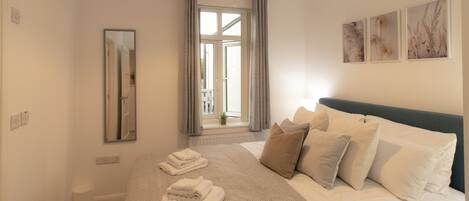 A neatly arranged bedroom with a large bed, a bedside table, a mirror, and a window with curtains.