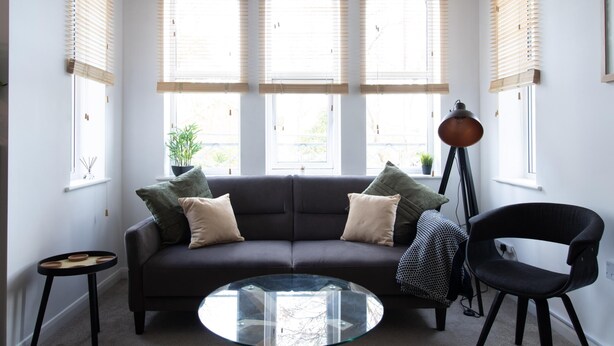 Een moderne woonkamer met een donkergrijze bank, drie ronde ramen met zonwering, een glazen salontafel, een zwarte vloerlamp en een klein bijzettafeltje.
