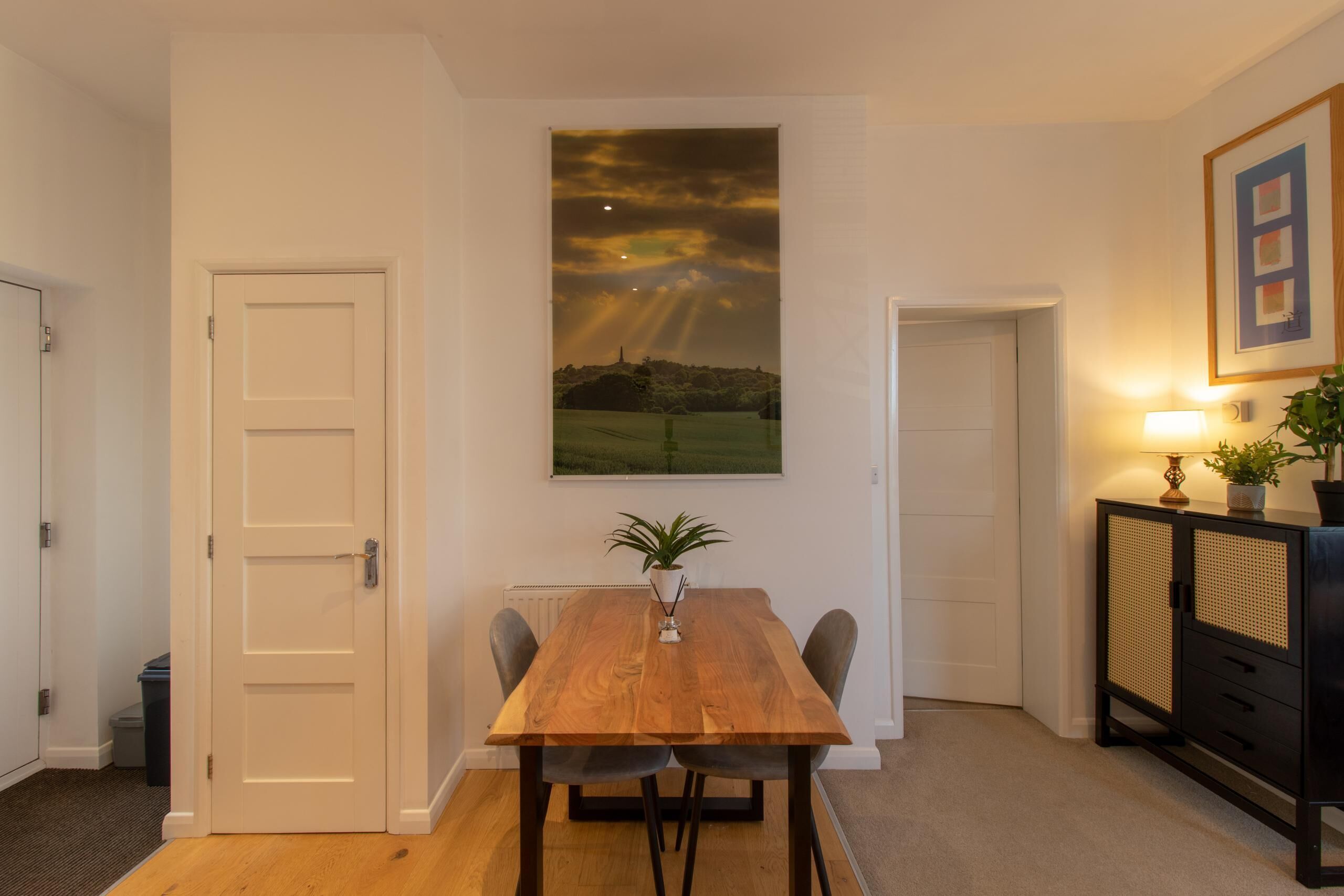 Uma sala de jantar com uma mesa de madeira, duas cadeiras, um vaso com planta e um quadro de uma paisagem.