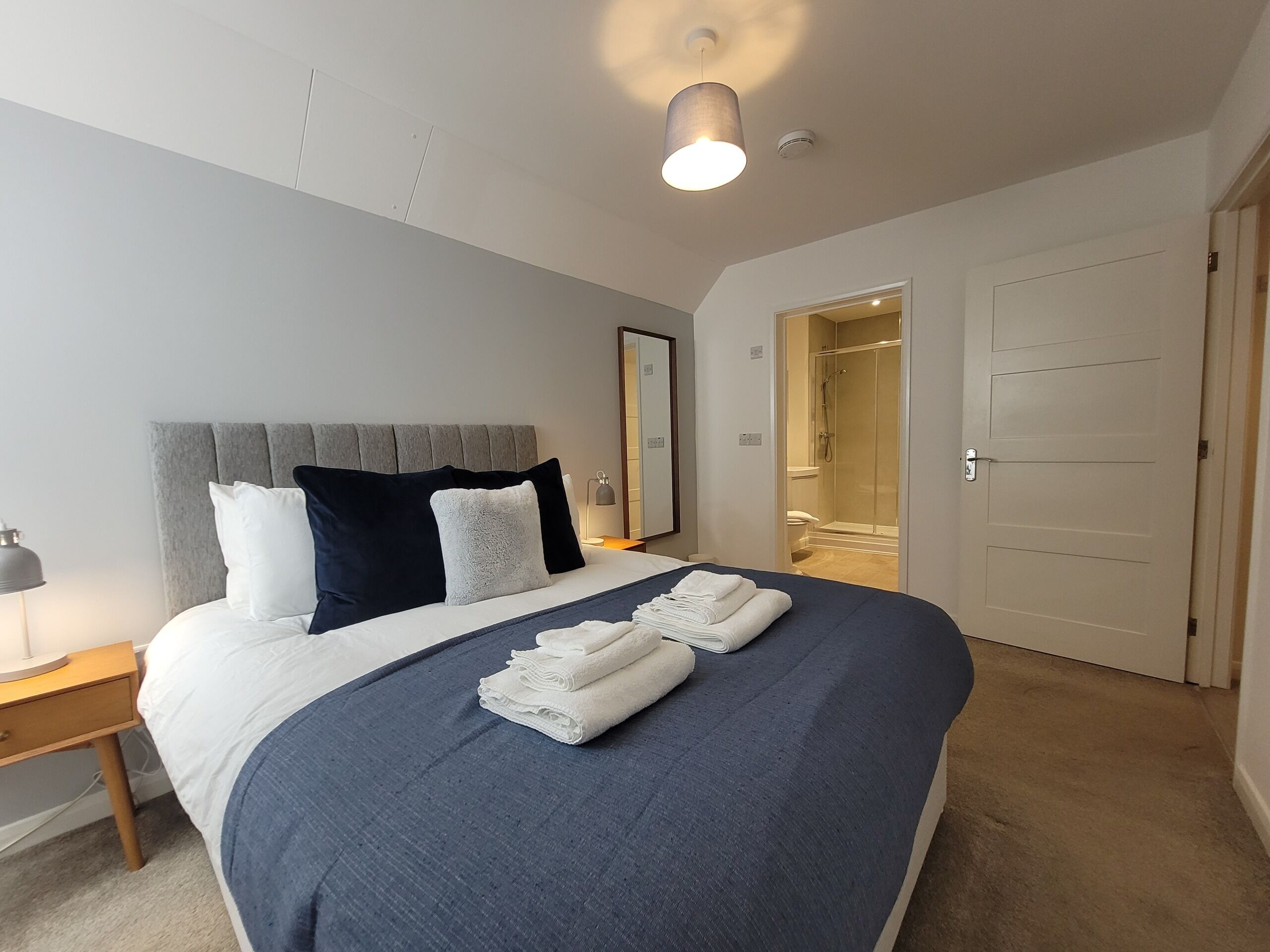 A neatly made bed with a grey headboard, a bedside lamp, and a stack of folded towels.