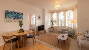Een moderne woonkamer met een houten eettafel, twee stoelen, een bank, een salontafel en een televisie. Er zijn grote ramen met luxaflex, een vloerlamp en een schilderij aan de muur.